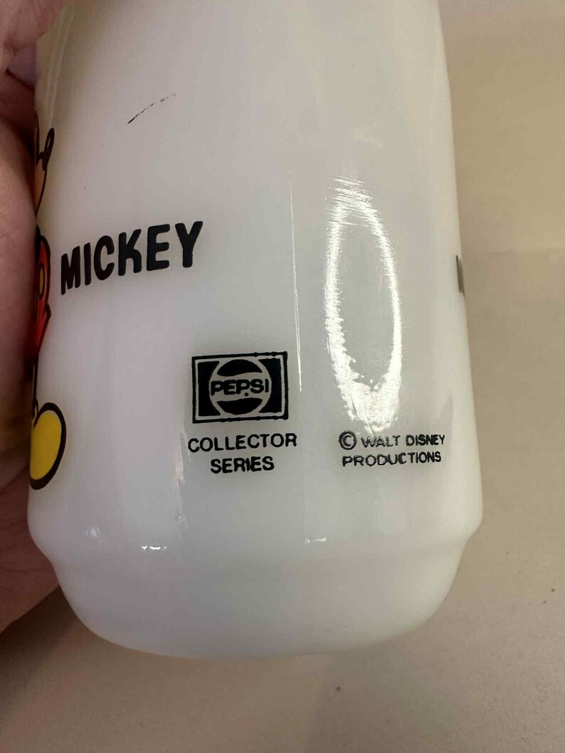 Pu&ograve; includere: Bicchiere bianco Pepsi Collector Series con grafica di Mickey Mouse. Il bicchiere presenta il logo Pepsi e il testo "Collector Series". Il testo "Mickey" &egrave; visibile in nero. Il fondo del bicchiere riporta la scritta "Walt Disney Productions".