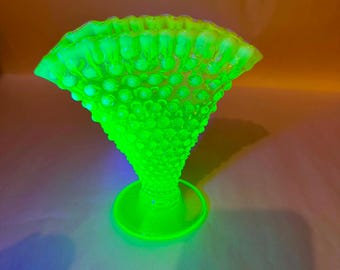 May include: A vibrant neon green glass vase featuring a textured hobnail pattern across its surface. The vase has a wide, flared, and ruffled rim that tapers down to a circular pedestal base. The glass glows with a bright, intense luminescence.