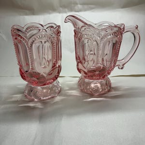 May include: A pink glass creamer and sugar bowl set. The creamer has a handle and spout, while the sugar bowl is open. Both pieces have a starburst design on the base and a scalloped rim.