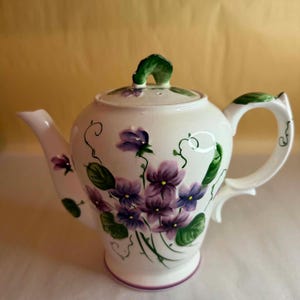 May include: A white ceramic teapot featuring a hand-painted floral design of purple violets with green leaves and vines. The teapot has a matching lid with a green handle, a curved spout, and a sturdy handle, all decorated with delicate painted vines.