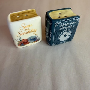 May include: Two ceramic book-shaped salt and pepper shakers. One is white with the text Sense and Sensibility and floral accents. The other is dark blue with the text Pride and Prejudice, a silhouette illustration, and white filigree details. Both have tan tops with holes.