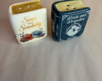 May include: Two ceramic book-shaped salt and pepper shakers. One is white with the text Sense and Sensibility and floral accents. The other is dark blue with the text Pride and Prejudice, a silhouette illustration, and white filigree details. Both have tan tops with holes.