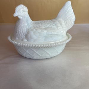 May include: A white hen-shaped covered dish. The hen sits atop a basket-weave base. The hen's body has a textured pattern, and the head and tail are detailed. The dish is made of opaque white glass.