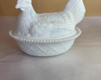 May include: A white hen-shaped covered dish. The hen sits atop a basket-weave base. The hen's body has a textured pattern, and the head and tail are detailed. The dish is made of opaque white glass.