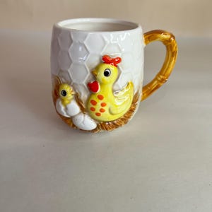 Può includere: Una tazza in ceramica bianca con un design di pollo e pulcino giallo. La tazza ha un motivo a nido d'ape e un manico marrone.