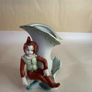 Könnte beinhalten: Eine Vintage-Keramikvase mit einer sitzenden Figur in roter und grüner Kleidung. Die Figur befindet sich neben einer weißen Vase mit einem einzigartigen, geschwungenen Design. Die Vase hat einen hellgrünen Rand und Blattakzente, was eine skurrile Ästhetik erzeugt.