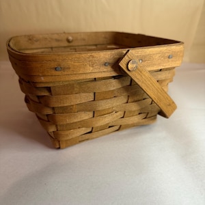 Puede incluir: Una cesta rectangular de madera tejida con asa. La cesta está hecha de madera de color marrón claro y tiene un asa sujeta con una clavija de madera. Los lados de la cesta están tejidos con un patrón entrecruzado.