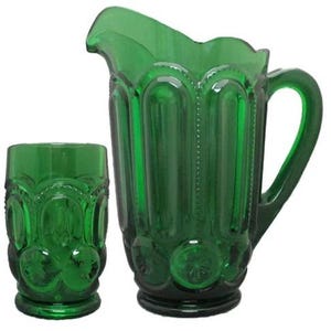 May include: A green glass jug and tumbler set. The jug has a handle and a scalloped rim. The tumbler is short and cylindrical. Both pieces have a textured, embossed design. The set is made of a deep emerald green glass.