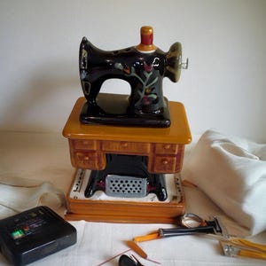 May include: A ceramic sewing machine figurine featuring a black upper machine body with floral accents, resting on a mustard yellow desk base with brown drawers and a black treadle stand. Nearby are a black tape measure and sewing tools.