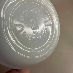 Pu&ograve; includere: Primo piano del fondo di una ciotola di colore chiaro, possibilmente in vetro. Il testo "OVEN-PROOF" &egrave; visibile, insieme a un simbolo di ancora e alle parole "MADE IN USA". La ciotola ha una forma arrotondata.