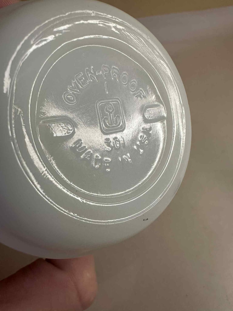 Pu&ograve; includere: Primo piano del fondo di una ciotola di colore chiaro, possibilmente in vetro. Il testo "OVEN-PROOF" &egrave; visibile, insieme a un simbolo di ancora e alle parole "MADE IN USA". La ciotola ha una forma arrotondata.