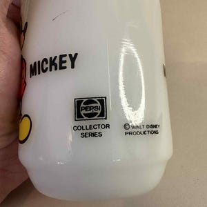 Pu&ograve; includere: Bottiglia bianca Pepsi Collector Series con grafica di Mickey Mouse. La bottiglia presenta la scritta "Mickey" in nero, un logo Pepsi e il testo "Walt Disney Productions".