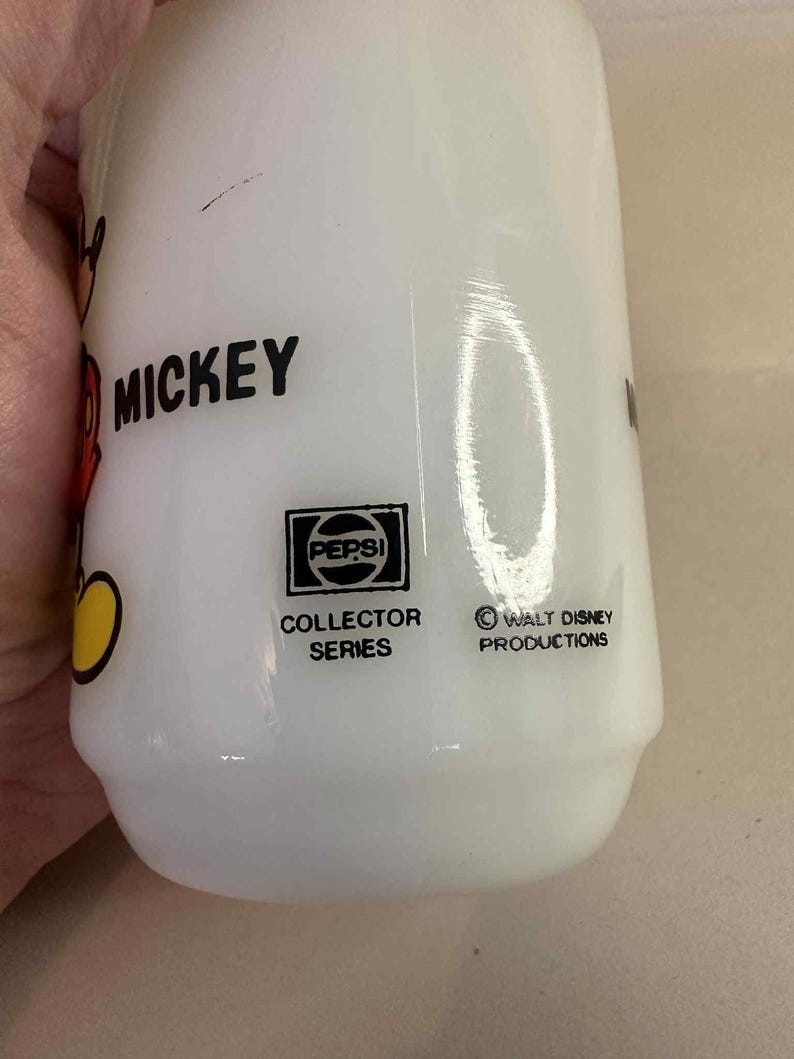 Pu&ograve; includere: Bottiglia bianca Pepsi Collector Series con grafica di Mickey Mouse. La bottiglia presenta la scritta "Mickey" in nero, un logo Pepsi e il testo "Walt Disney Productions".