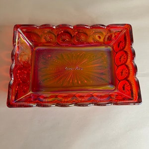 May include: A red glass dish with a scalloped edge and a starburst pattern. The dish has a clear iridescent finish and is decorated with a raised design of stars and circles.