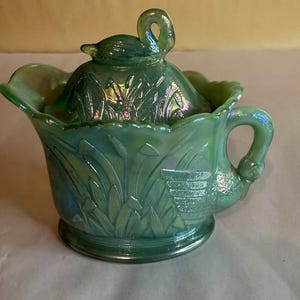 Puede incluir: Un plato de vidrio verde iridiscente con forma de cisne con tapa. El plato presenta un asa de cisne y un asa de tapa con forma de cisne. El diseño incluye juncos en relieve y una cesta. El plato tiene aproximadamente 15 cm de diámetro.