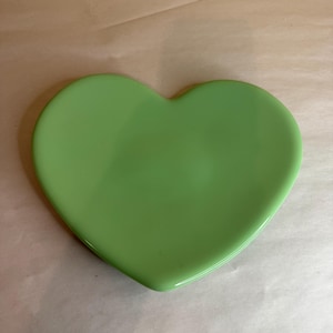 Op de afbeelding: Een hartvormige, lichtgroene keramische plaat. De plaat heeft een gladde, glanzende afwerking en is gecentreerd in het frame. De plaat heeft een effen, levendige groene kleur.