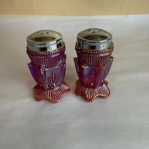 以下が含まれることがあります： 虹色のガラス製ソルト＆ペッパーシェーカーのペア。シェーカーは、フレアベースとシルバーメタルトップを備えた、ふっくらとした装飾的なデザインです。ガラスは、オレンジ色のハイライトのある赤紫色の色合いです。