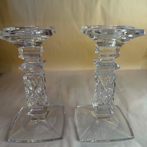 Puede incluir: Dos candelabros de cristal transparente. Cada uno tiene una base cuadrada, un tallo con diseño de diamantes y una parte superior circular para una vela. Los candelabros son de vidrio transparente y reflejan la luz.