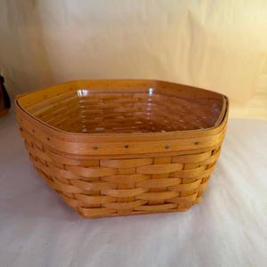 Puede incluir: Una cesta hexagonal tejida con tiras de madera de color marrón claro. La cesta presenta un borde de madera maciza lisa asegurado con pequeños clavos de metal a lo largo del borde superior. Los lados tienen un patrón de tejido clásico.