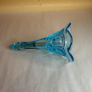 Könnte beinhalten: Eine lichtblaue, durchscheinende Glasvase mit einer trompetenförmigen Öffnung und einem gewellten, gebogten Rand. Der Körper der Vase weist ein strukturiertes, geprägtes Muster auf und verjüngt sich zu einem schmalen, zylindrischen Fuß.
