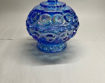 Puede incluir: Un azucarero de vidrio azul iridiscente con cuerpo redondo y tapa abovedada. El plato tiene un diseño en relieve de pequeños círculos.