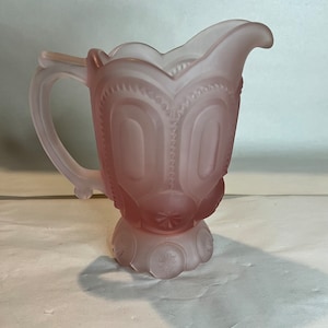 May include: A pink frosted glass creamer with a handle and spout. The creamer features an embossed design of arches and starbursts. The base has a decorative starburst pattern. The creamer is a vintage style.