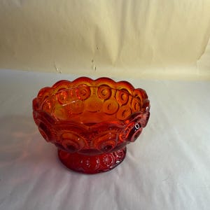Könnte beinhalten: Eine leuchtend rote Glasschale mit einem sich wiederholenden Mond- und Sternmuster, das außen eingeprägt ist. Die Schale hat einen gewellten Rand und steht auf einem runden Sockelfuß, mit einer durchscheinenden Oberfläche.