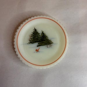 May include: A small, round, white plate with a scalloped edge and a painted winter scene. The scene features two green pine trees, a red bird, and a snowy ground. A thin orange line borders the inner edge of the plate.