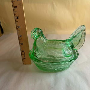 Może przedstawiać: Zielona szklana miska w kształcie kury z pokrywką, osadzona w podstawie przypominającej gniazdo. Miska ma około 15 cm długości, z teksturowaną powierzchnią. Pokrywka ma kształt kury ze szczegółowymi wzorami piór.