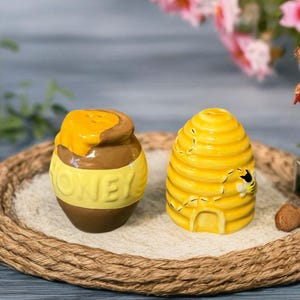 Könnte beinhalten: Ein Keramik-Salz- und Pfefferstreuer-Set, bestehend aus einem Honigtopf und einem Bienenkorb. Der Honigtopf ist braun mit einem gelben Band mit der Aufschrift "HONEY". Der Bienenkorb ist gelb mit einer kleinen Biene und einer Tür.