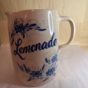 May include: Cream-colored ceramic pitcher with a curved handle and spout. The word "Lemonade" is written in blue script across the front. Blue floral and swirl designs decorate the pitcher, creating a vintage aesthetic.