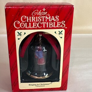 May include: A red box with a white label that reads "Gibson Christmas Collectibles" and features a small bell ornament inside. The bell is clear glass with a gold rim and a red ribbon. The ornament features a small figurine of a woman in a red dress and blue shawl. The text "Ringing in Christmas Ornament" is printed on the box.