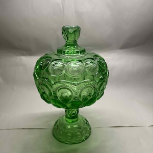 May include: A green glass candy dish with a lid. The dish is round with a pedestal base and a decorative pattern of stars and moons. The lid has a small handle. The dish is approximately 8 inches tall.