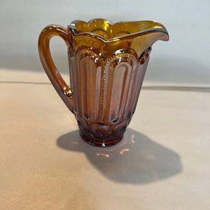 Op de afbeelding: Een kleine kan van amberkleurig glas met een gegolfde rand en een handvat. De kan heeft een textuur met verticale lijnen en kleine cirkels.