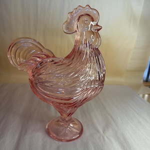 May include: A pink glass rooster figurine with a textured, swirled design. The rooster stands on a pedestal base and features a detailed comb, wattle, and tail feathers. The glass has a translucent quality.
