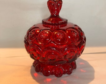 Puede incluir: Un bombonera de cristal rojo rubí con tapa. El plato es redondo con un patrón de estrella y una base de pedestal. La tapa tiene un asa en forma de corazón. El objeto es probablemente vintage.