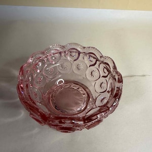 Könnte beinhalten: Eine rosa Glasschale mit gewelltem Rand und kreisförmigem Boden. Die Außenseite der Schale weist ein sich wiederholendes Muster aus Kreisen und Sternen auf. Die Schale besteht aus transparentem rosa Glas.
