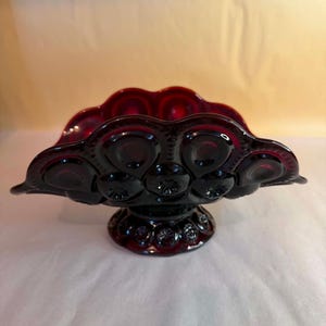 May include: A deep red glass bowl featuring a scalloped rim and a repeating moon and star pattern across its exterior surface. The bowl is supported by a circular pedestal base that also displays the same embossed moon and star design.
