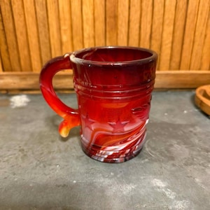 May include: A red glass mug with a handle. The mug has a star pattern and a swirled white and red design. The mug is approximately 10 cm tall.