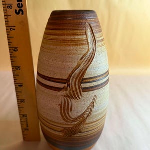 May include: A handmade ceramic vase with a textured surface and a unique, carved design. The vase is primarily beige and brown, with horizontal stripes and a dark brown rim. The vase measures approximately 7 inches in height.