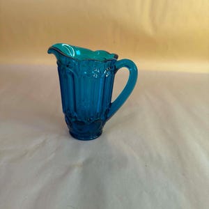 Op de afbeelding: Een kleine roomkan van doorschijnend blauwgroen glas met een geschulpte rand, een gebogen handvat en een verticaal geribbeld patroon op het lichaam. De voet van de kan is versierd met een reeks in reliëf aangebrachte cirkelvormige motieven.