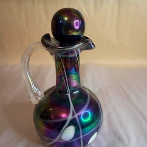 Puede incluir: Decantador de vidrio iridiscente con tapón esférico. El decantador tiene un cuerpo redondo, un borde acampanado y un asa transparente. Líneas y acentos blancos decoran el cuerpo. Los colores cambian de morado a verde y azul.