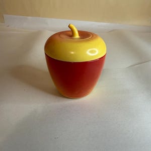 May include: A ceramic apple-shaped container with a yellow top and a red base. The container has a stem-like handle on the top. The apple-shaped container is likely used for storage or decoration.