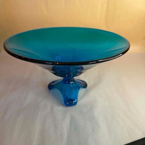 May include: A vibrant turquoise blue glass bowl with a wide, shallow basin and a smooth, reflective surface. The bowl is supported by a sturdy, three-legged pedestal base, creating a balanced and elevated silhouette for this decorative glass piece.