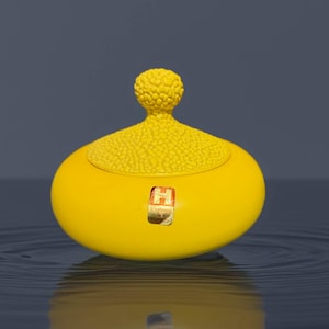 以下が含まれることがあります： 明るい黄色のセラミックジャー。テクスチャード加工の蓋と小さな丸いノブが付いています。ジャーは滑らかな楕円形で、前面に小さな長方形のステッカーが付いています。ジャーは水面に反射しています。