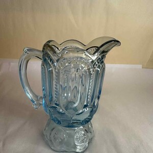 May include: A light blue glass jug with a handle and a decorative base. The jug has a scalloped rim and is adorned with starburst and floral patterns. The jug is approximately 15 cm tall.