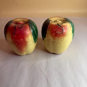 May include: A set of two ceramic salt and pepper shakers shaped like apples. The shakers are painted with red, yellow, and green colors. The word "P" is painted on one shaker and the word "S" is painted on the other shaker.