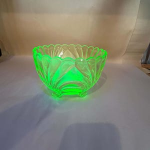 Può includere: Una ciotola in vetro verde neon vibrante con bordo smerlato e un design esterno a coste simile a petali. La ciotola è sostenuta da tre piccoli piedini integrati alla base, che mostrano un aspetto traslucido e luminoso sotto la luce.