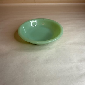 Könnte beinhalten: Eine kleine, runde Schüssel aus jadegrünem Glas. Die Schüssel hat eine geringe Tiefe und eine glatte, glänzende Oberfläche. Die Farbe ist ein weiches, gedämpftes Grün, und die Schüssel scheint leer zu sein.