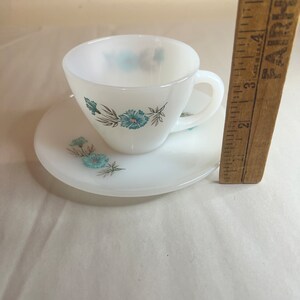 May include: A white teacup and saucer set with a floral design featuring blue flowers and green leaves.
